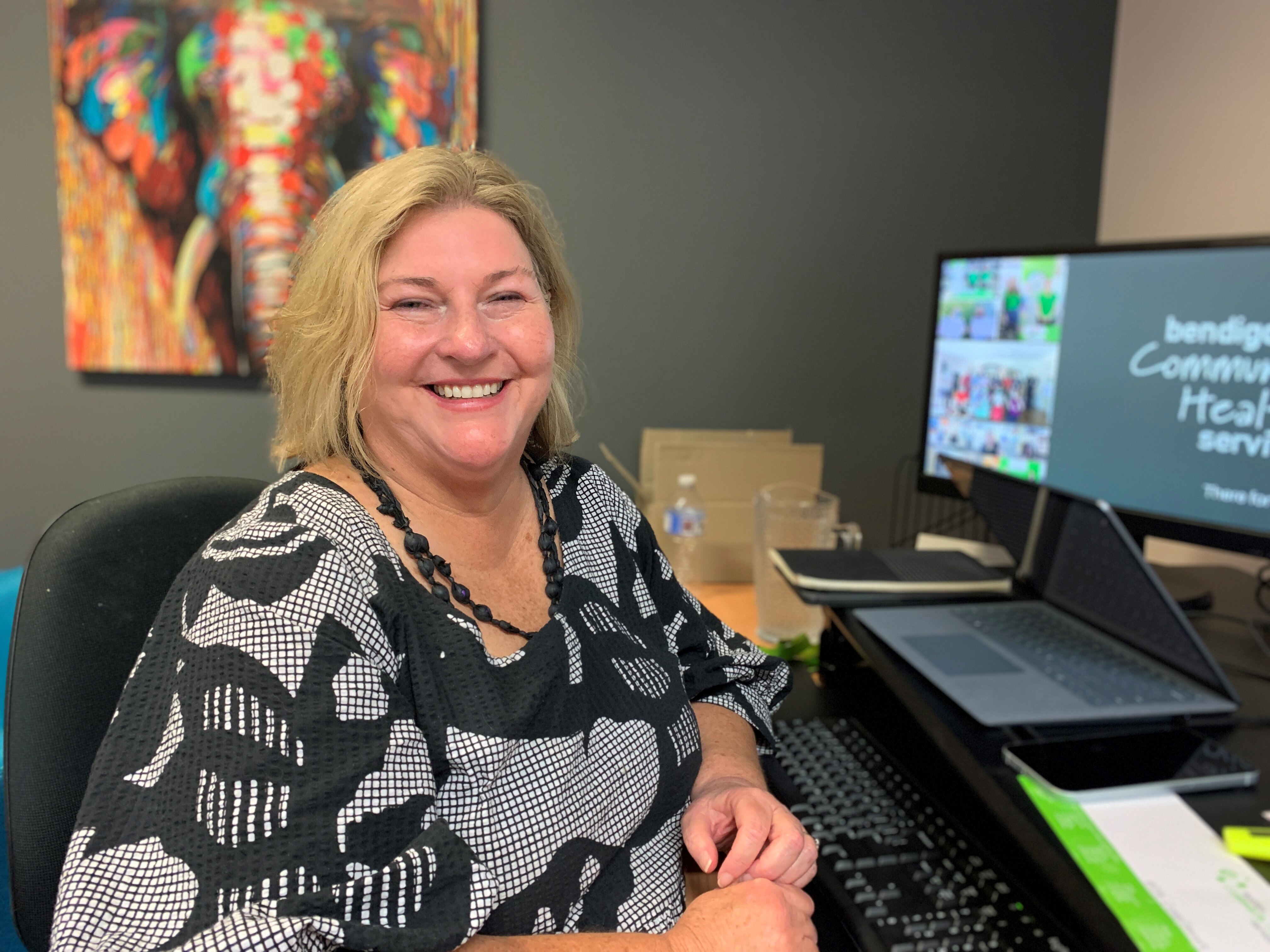 A smiling woman with a blonde bob haircut, wearing a black and white shirt sitting at a desk.