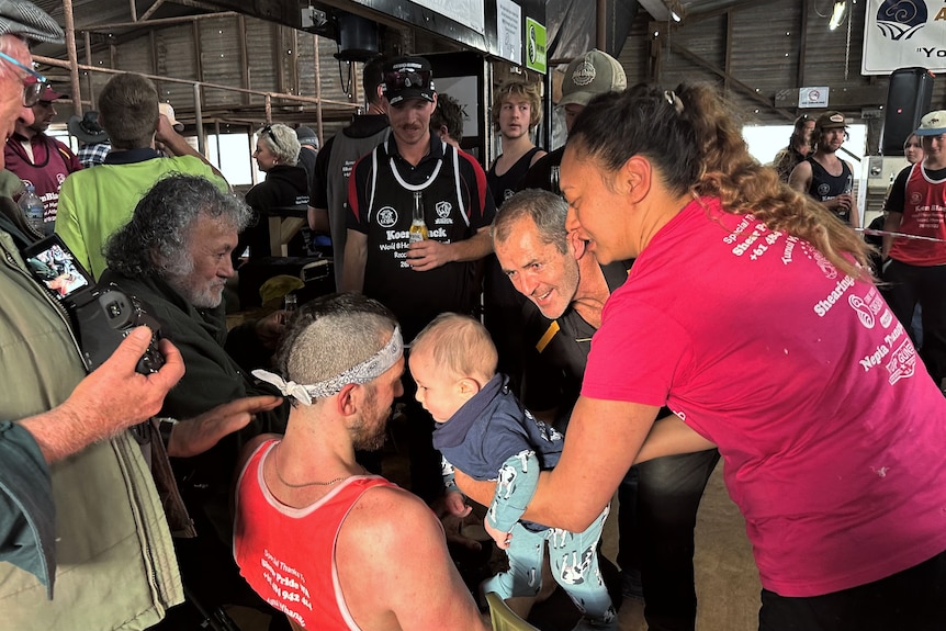 A man wearing a red singlet is surrounded by smiling family and friends in a shearing shed