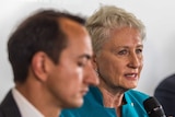 a woman speaks with a microphone with a man out of focus in the foreground