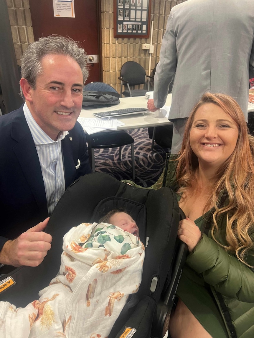 A man in a suit and a woman with long hair smile at the camera with a baby in a capsule between them.