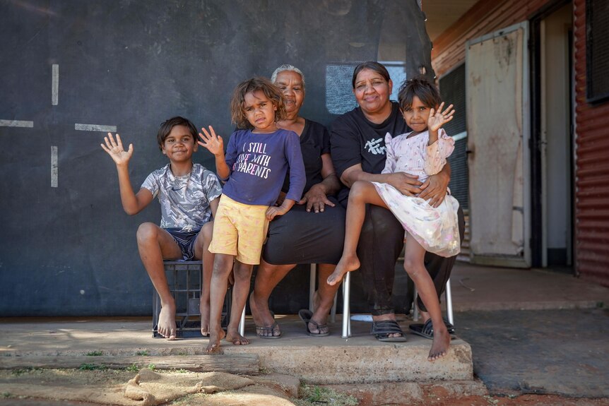A group of two Aboriginal women and three kids sit together in front of a house