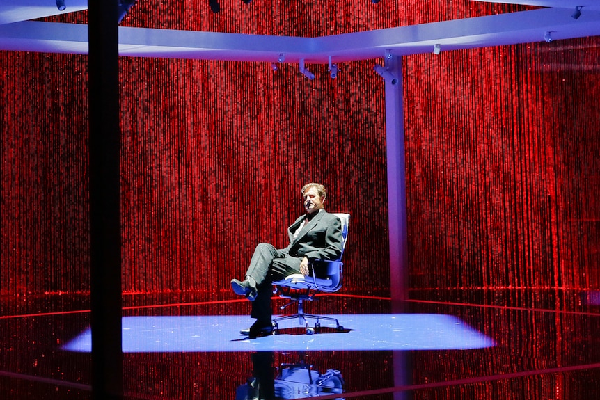 Darkened stage with backdrop curtain of red LED lights and and man sitting in chair under a blue-lit square pavilion structure.