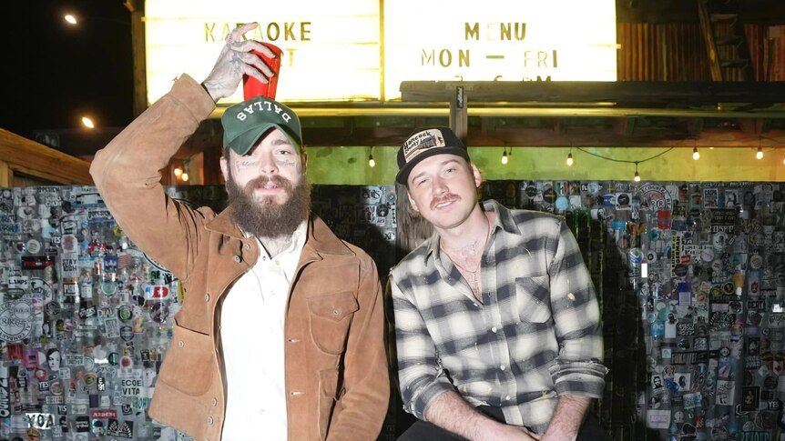 post malone and morgan wallen sit next to each other in a dive bar. post holds a red cup on top of his head.