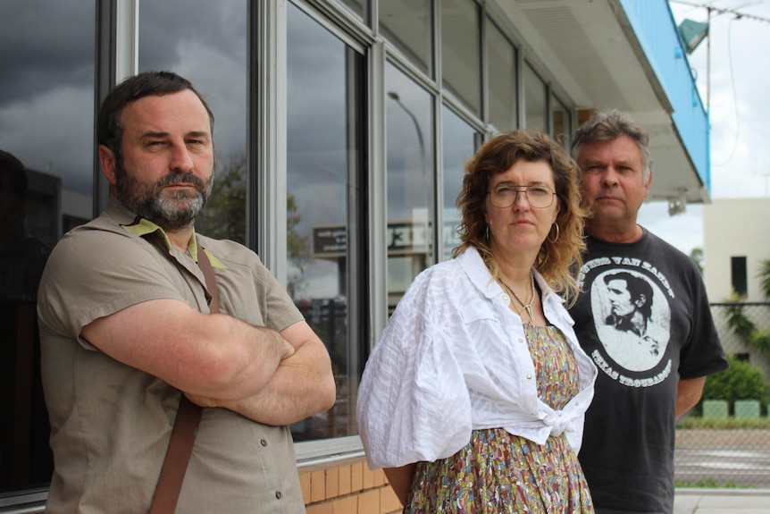 Three people standing in front of a brick building looking upset.