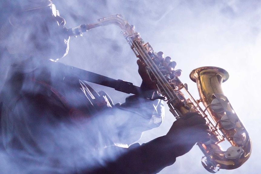 Surrounded by light smoke, a man wearing beret and glasses plays a saxophone, holding it upwards.