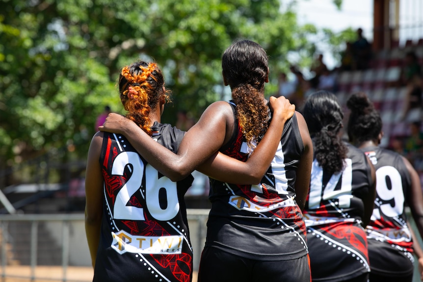 Two women in red and black AFL guernseys hold each other arm in arm