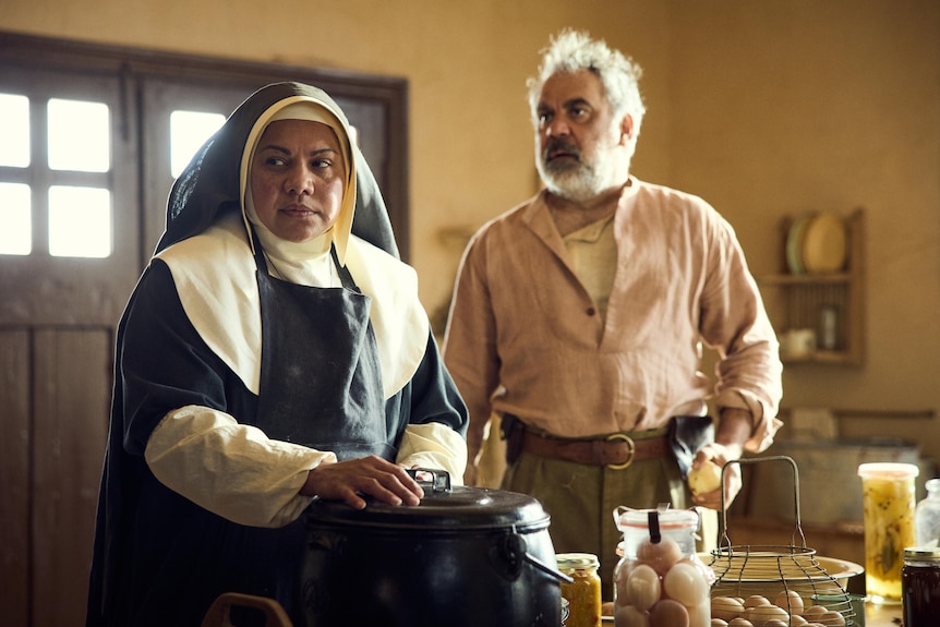 An Aboriginal woman dressed as a nun, standing in a country kitchen. An Aboriginal man stands behind her.