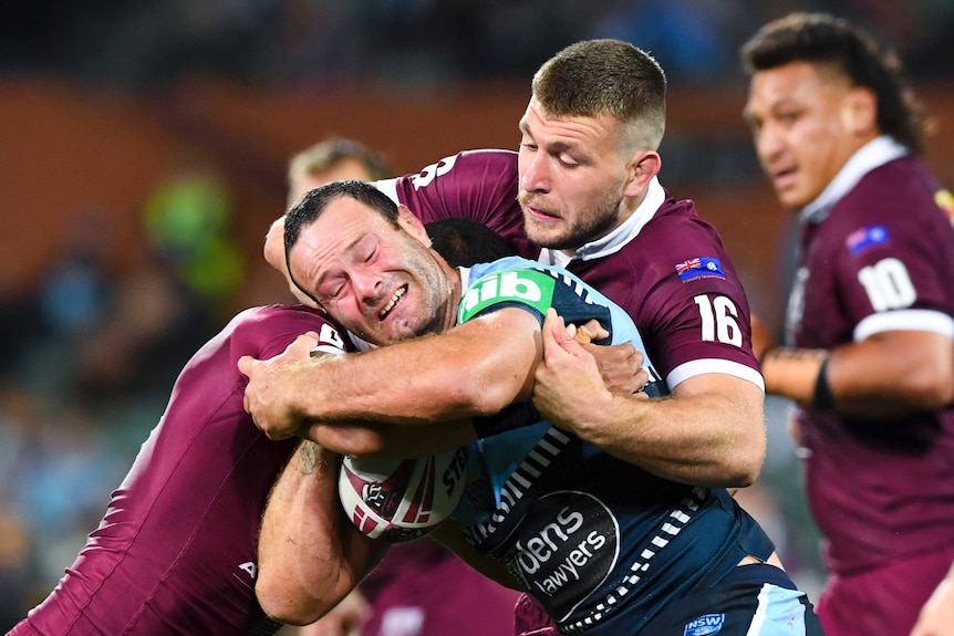 An NSW striker grimaces as he is held up in a tackle by an opposition striker in State of Origin.