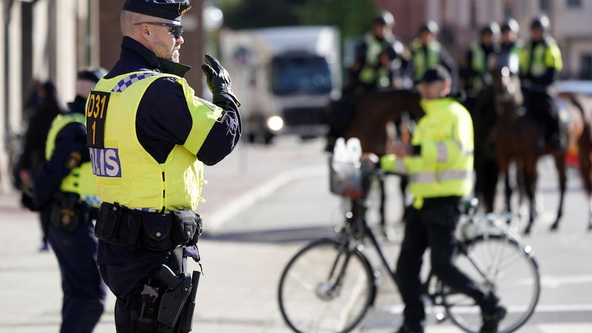 Police in high vis stand on the street as one in the background pushes a bike. 