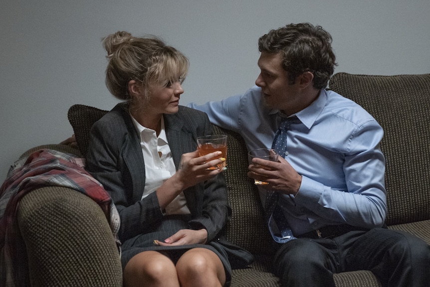 Young woman and man in office work clothes sitting on drab couch close together holding drinks, talking.