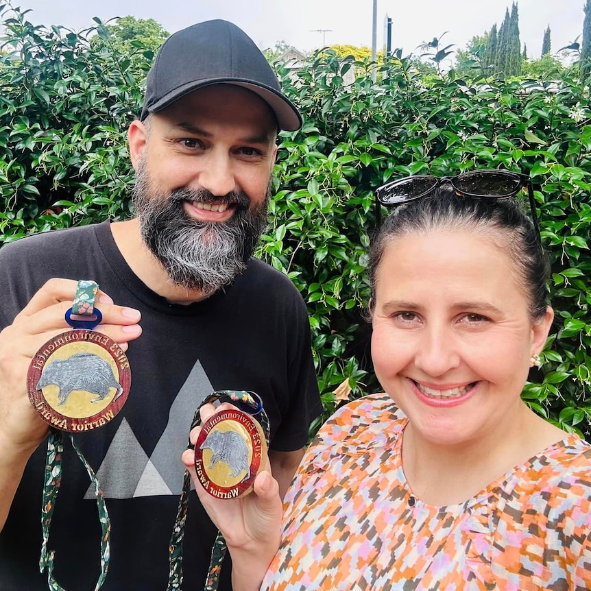 man with bear in black cap next to woman in patterned top with sunglasses on her head both holding medals on lanyards