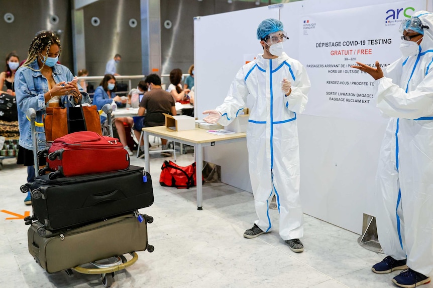 A woman with a trolley full of suitcases looks at her phone near two health workers in full PPE