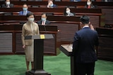 A man wearing a suit stands in front of Carrie Lam and raises his hand in oath.