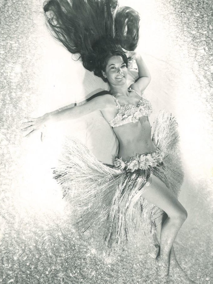 A black and white photo of a woman in a hula dancing costume lying on the floor with her hair fanned out