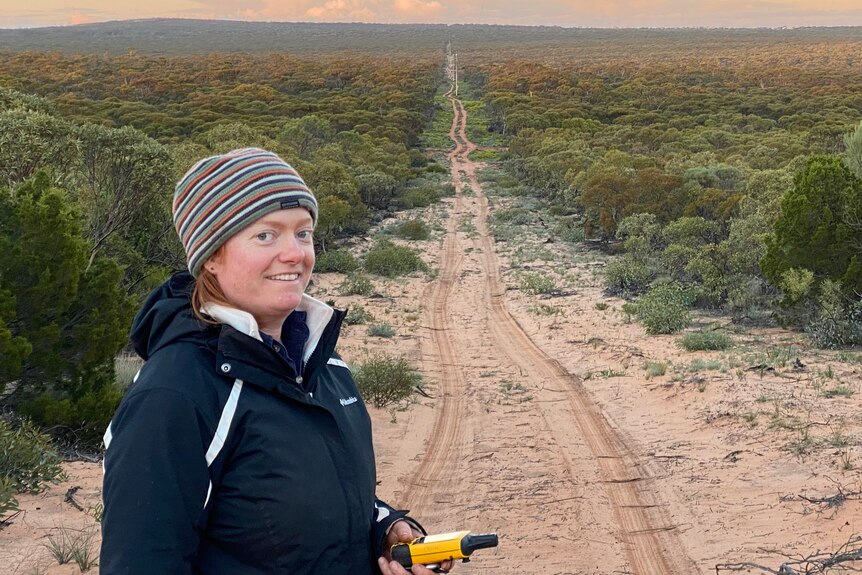 Woman on left looking at camera with long dirt road leading into distance with sunrise, green bush on either side