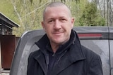 Michael O'Neill in a black jacket standing behind a van outside a house.