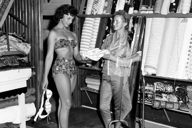 black and white photo of a woman in a shop with rolls of fabric holding fabric to a woman in a bikini in front of a mirror