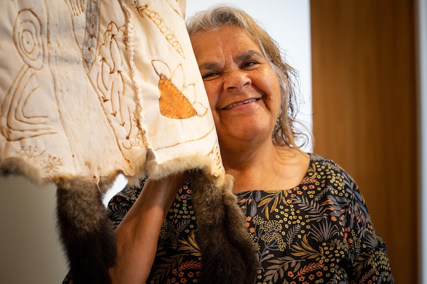 Loretta Parsley holds the underside of a possum skin cloak to her cheek, and looks at the camera smiling.