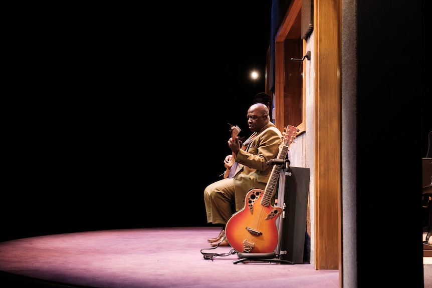 A bald African man plays guitar in a seated position, a second guitar resting in a stand beside him