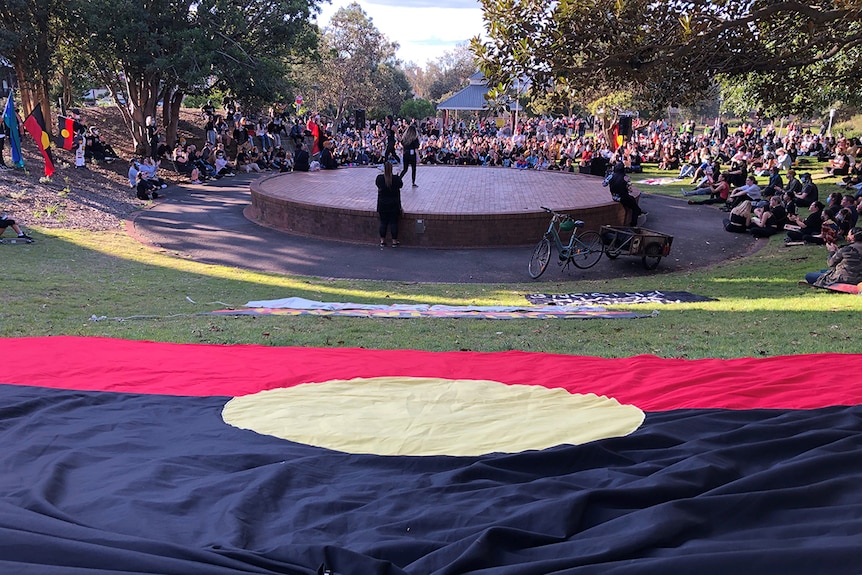 A large Indigenous flag is laid out on grass behind a rotunda where speaker address a large crowd.