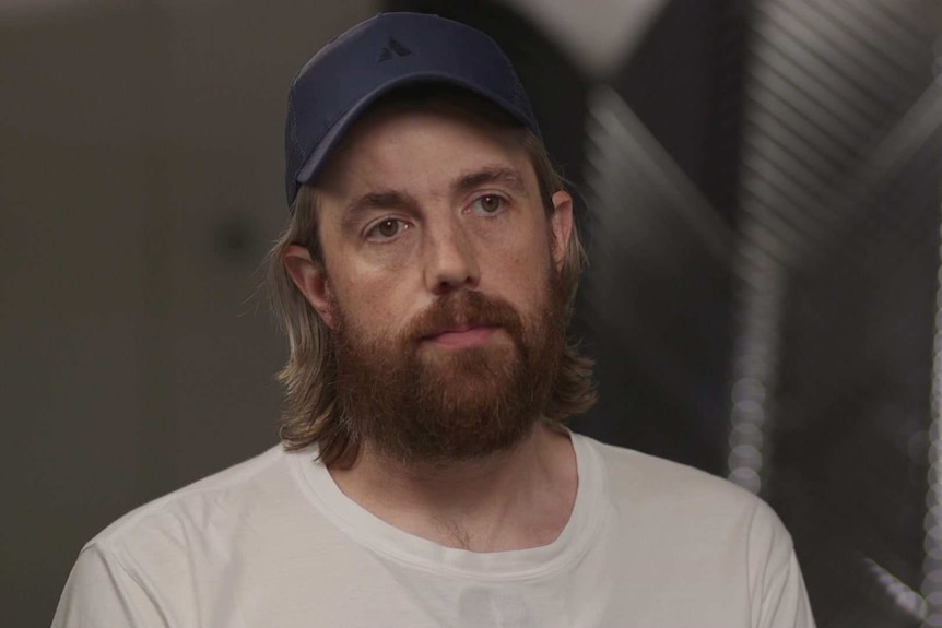 A young entrepreneur with long hair and a beard, wearing a cap while he appears on television.