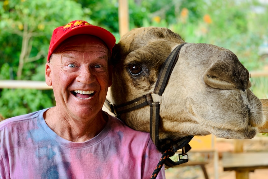 A man in a red cap with a surprised expression stands next to a camel
