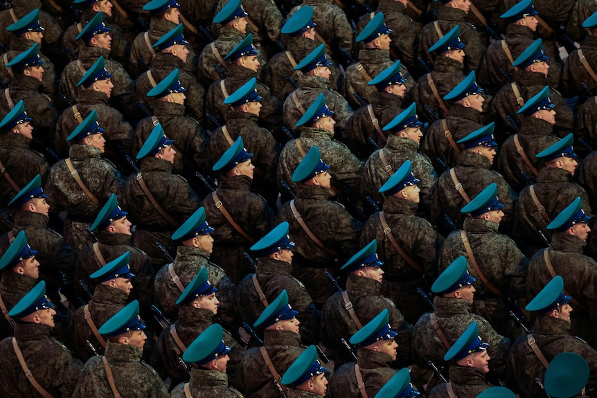 A group of soldiers stand in formation wearing miltiary uniforms and blue hats.