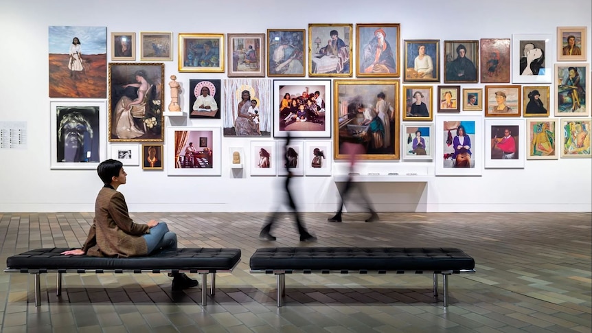 A woman sits on a bench inside a gallery, looking at a wall of portraits by women artists.