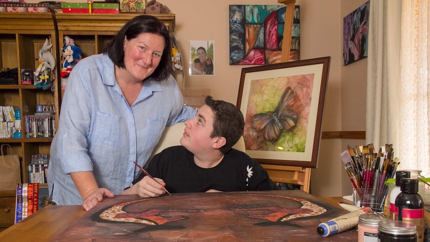 A woman leans over a boy painting an artwork at a table