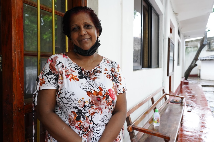 A middle-aged woman in white top with a floral pattern stands smiling next to a wooden door