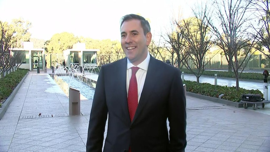 Treasurer Jim Chalmers smiles in a suit with a red tie standing outside Parliament. 
