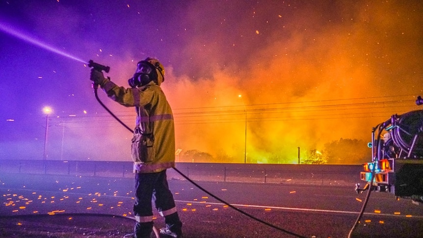DFES firefighter holds a hose as embers fall around him, fire in background