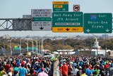 A big crowd runs on a major road in New York with signs above them to JFK Airport and other roads.