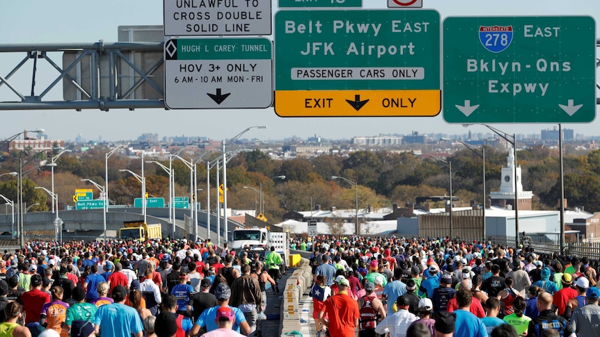 A big crowd runs on a major road in New York with signs above them to JFK Airport and other roads.