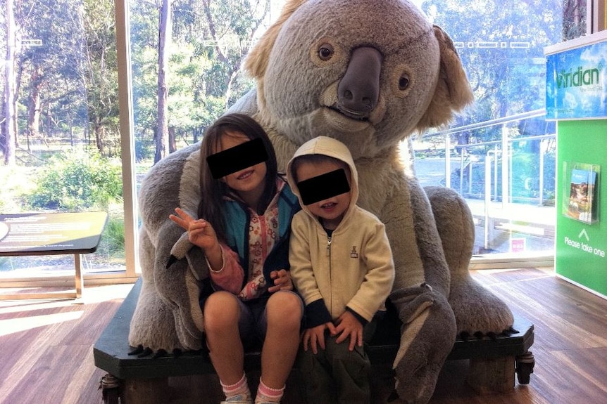 Two children sit on a toy koala, with anonymising black bars across their eyes.