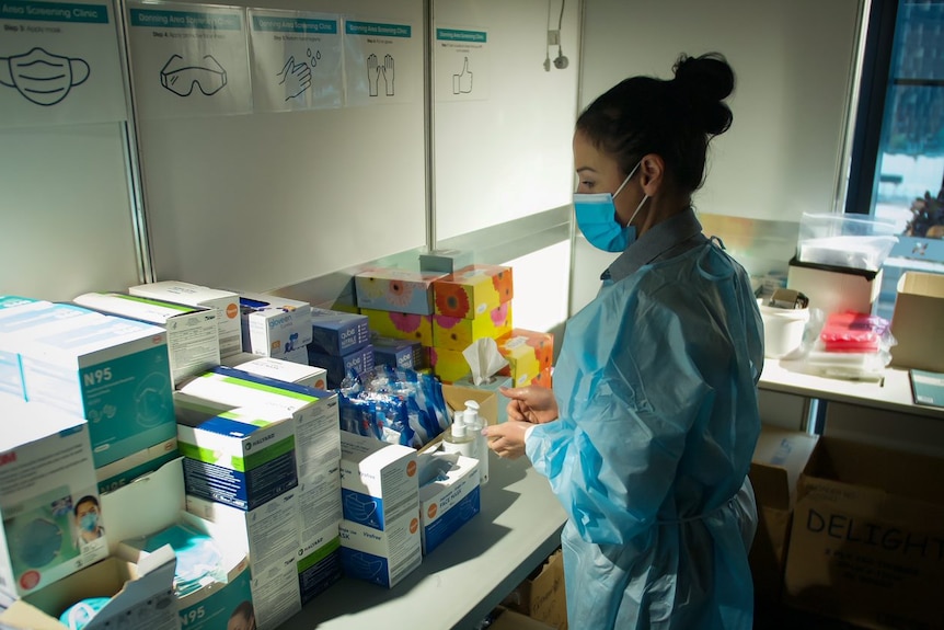 A woman wearing a mask and gown stands in front of boxes of medical supplies.