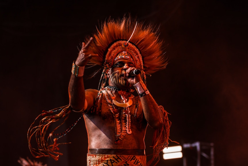 Man of PNG cultural heritage is dressed in traditional attire holds a mic on stage rapping. 