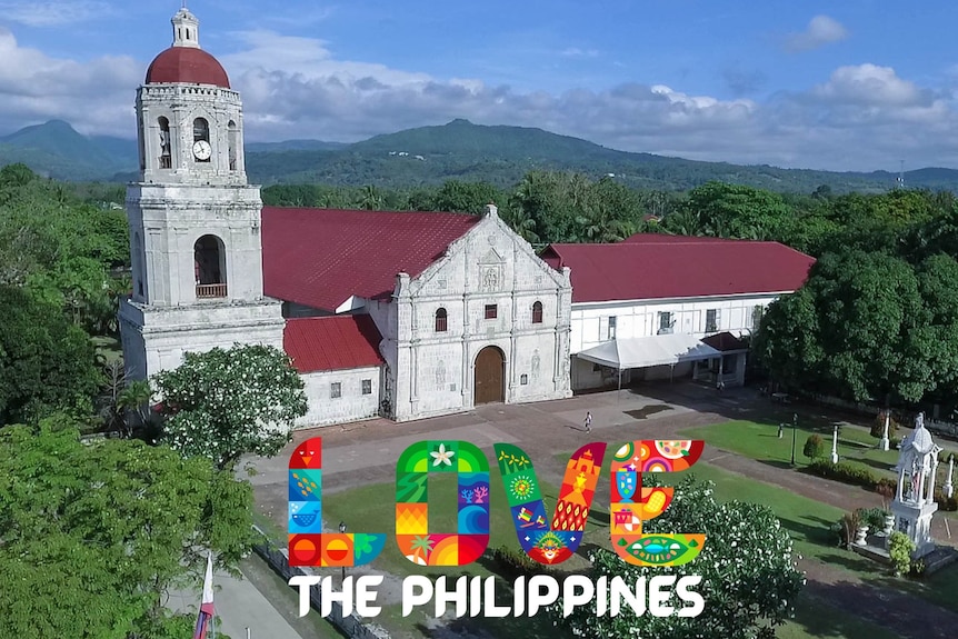 A white building with red roof and clock tower surrounded by green tress with colourful 'Love the Philippines' logo.