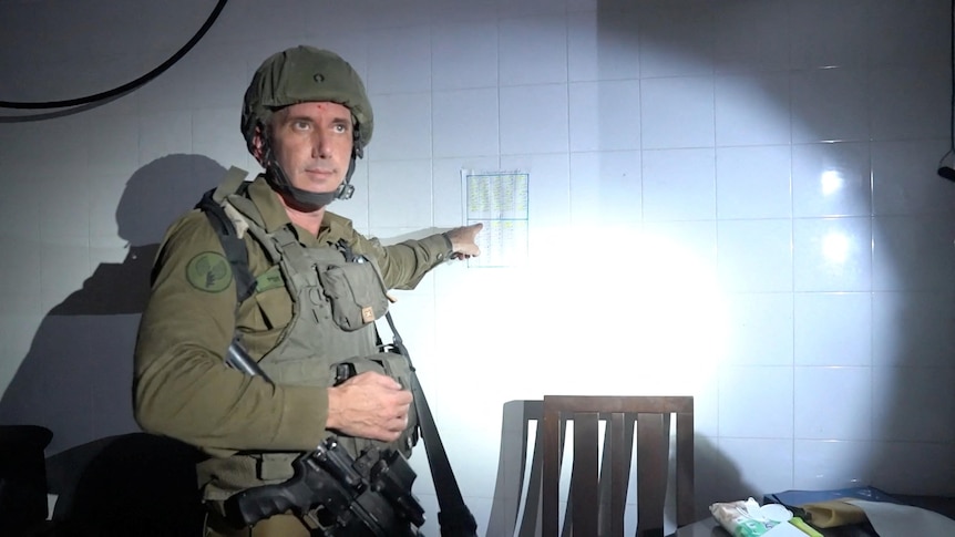 A man wearing military garb with weapons pointing at a piece of paper on the wall.