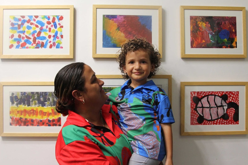 A woman holds a little girl in front of a wall of children's artworks.