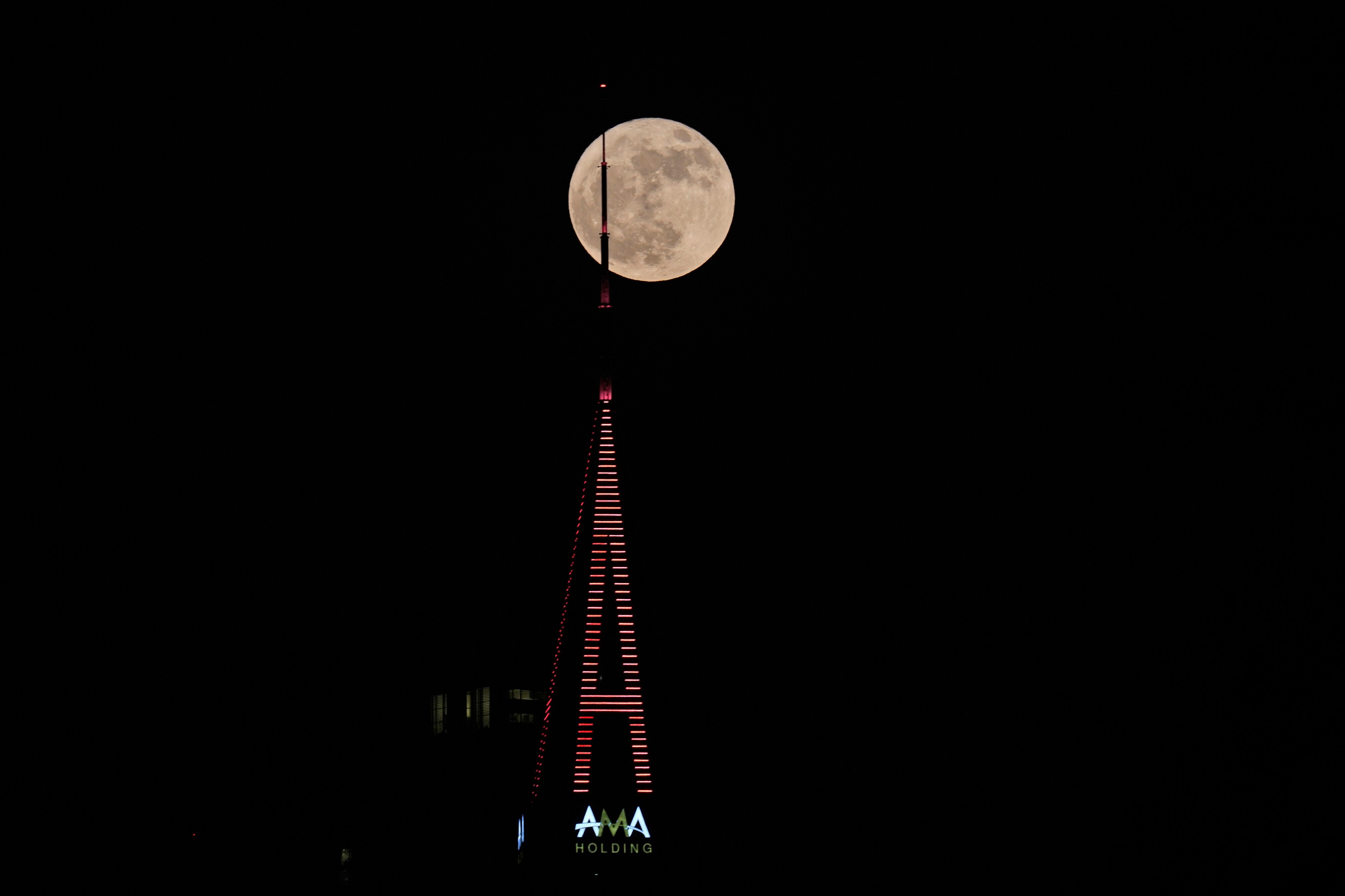 A large moon is seen behind the spire on top of a tall, thin tower lit with horizontal stripes of light.