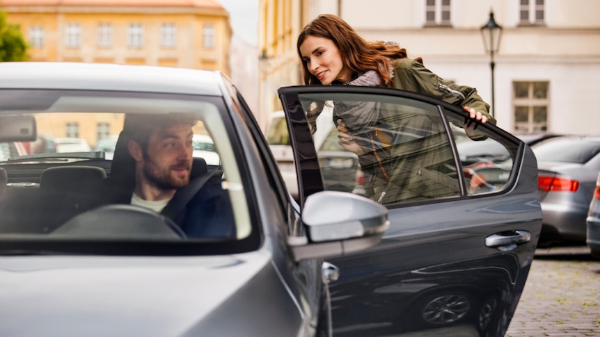 A lady goes to get into the back of a car as the driver looks in he side mirror