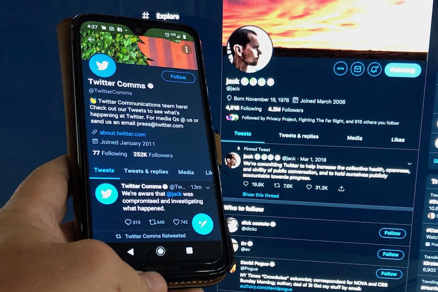 A hand holds a phone displaying Twitter Comms' tweet alongside a PC monitor displaying Jack Dorsey's Twitter account