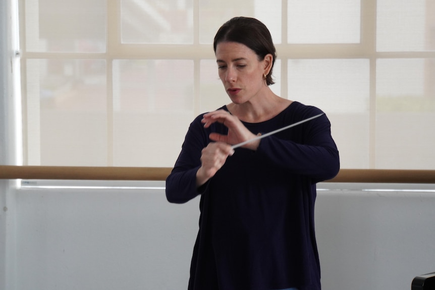 A woman holding a conducting baton looks down.