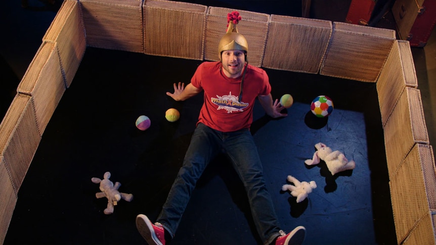 Man wearing helmet sits in pen with soft toys on floor