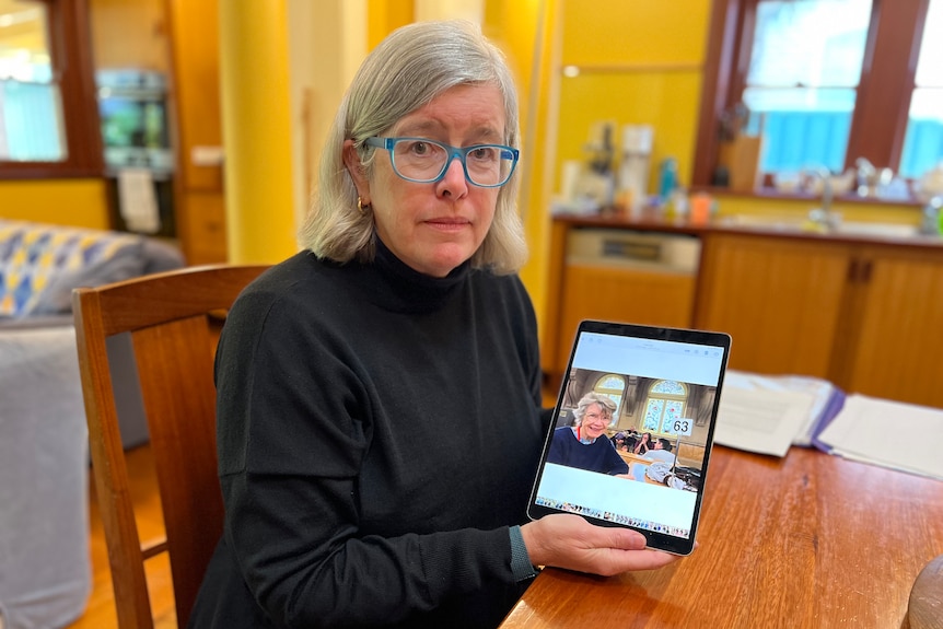 a woman wearing glasses sitting down and holding an ipad showing a picture of an elderly woman 