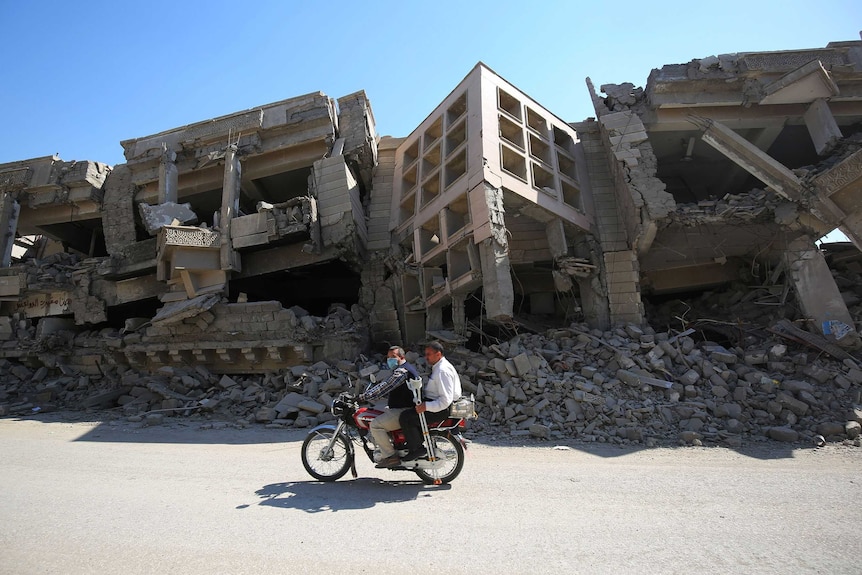 Two men on a motorbike ride past destroyed buildings.