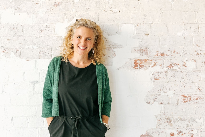 A smiling white woman with curly blonde hair wearing black clothes and a green cardigan stands against a whitewashed brick wall