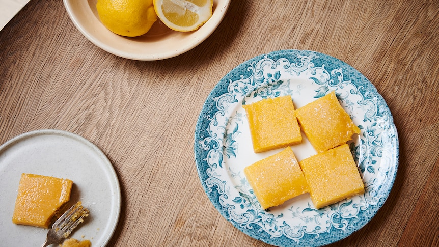 A plate of four lemon bars with a bowl of cut lemons in the background. A lemon bar is on a plate with a serving fork.