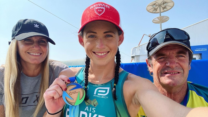 An Australian female athlete holds a World Triathlon Para Championships gold medal while next to two supporters.